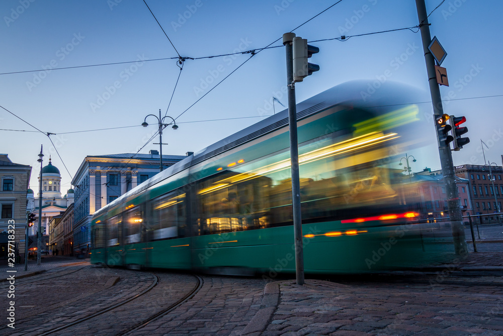 Public transportation in a city. Tram leaving the station, traffic lights, people, street, houses, overhead wires, rails. Movement and lights, long exposure. Public transportation in a city. Tram leaving the station, traffic lights, people, street, houses, overhead wires, rails. Movement and lights, long exposure.
