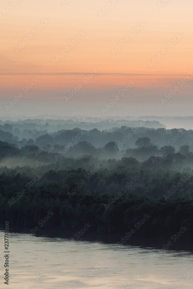 Mystical view on riverbank  of large island with forest under haze at early morning. Eerie mist among layers from tree silhouettes under predawn sky. Morning atmospheric landscape of majestic nature.