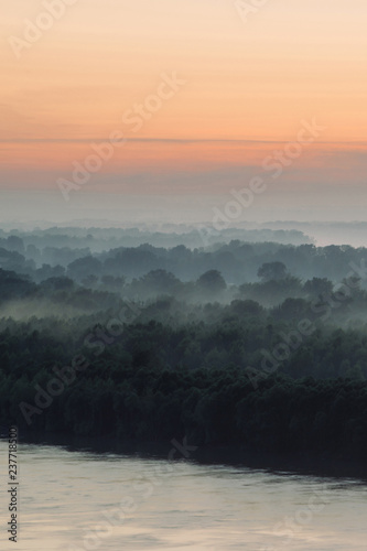 Mystical view on riverbank of large island with forest under haze at early morning. Eerie mist among layers from tree silhouettes under predawn sky. Morning atmospheric landscape of majestic nature.