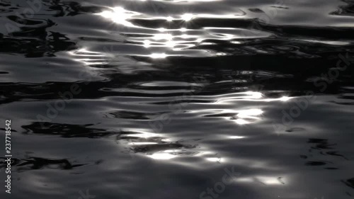 Slow motion of strip waving black water surface in sun shine reflection. Amazing abstract natural contrast background. High speed camera shooting. Bright play of light on calm ripple.
