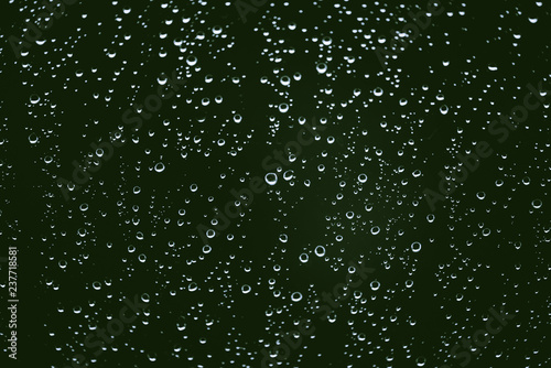 Dirty window glass with drops of rain. Atmospheric green background with raindrops. Droplets and stains close up. Detailed transparent texture in macro with copy space. Rainy weather.