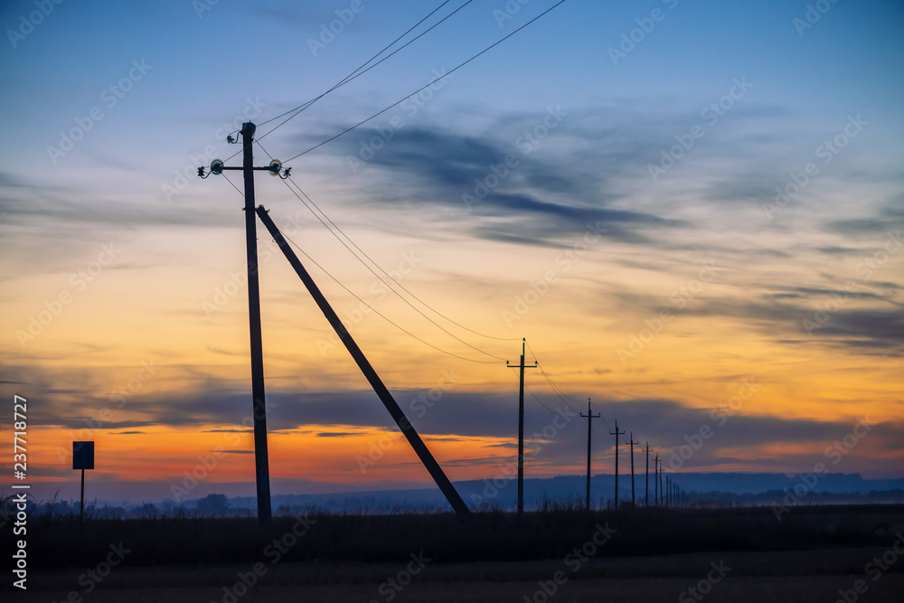 Power lines in field on sunrise background. Silhouettes of poles with wires at dawn. Cables of high voltage on warm orange blue sky. Power industry at sunset. Multicolored picturesque vivid sky.