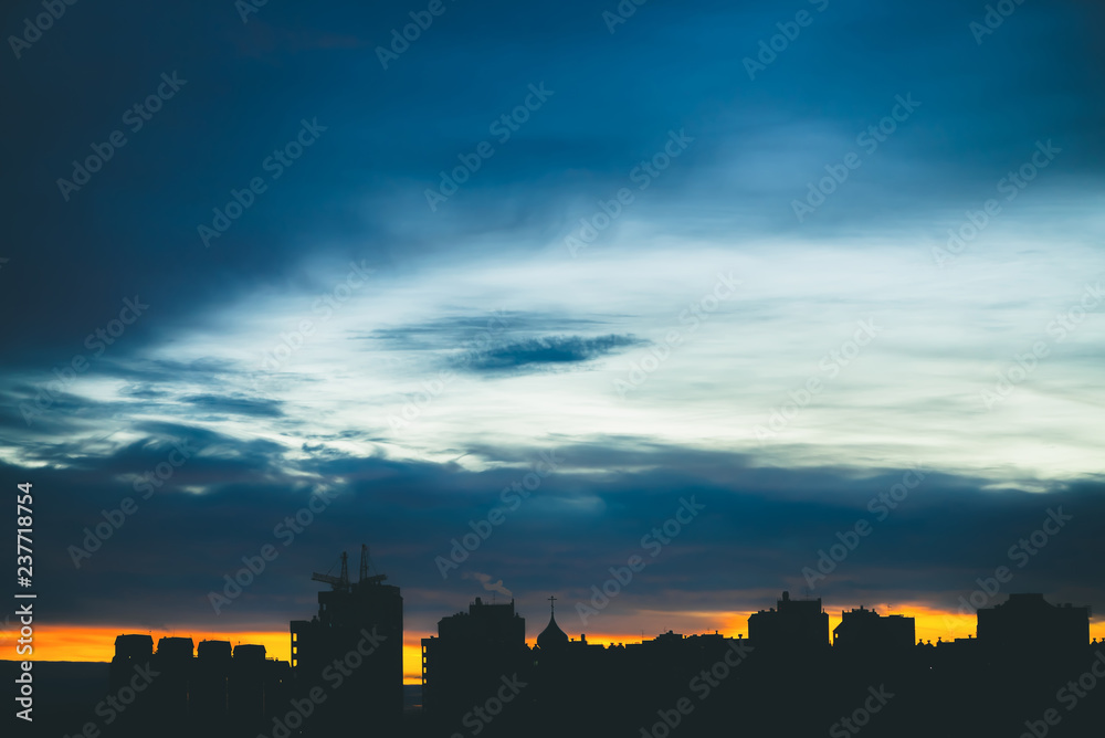Cityscape with wonderful varicolored vivid dawn. Amazing dramatic blue cloudy sky above dark silhouettes of city buildings. Atmospheric background of orange sunrise in overcast weather. Copy space.