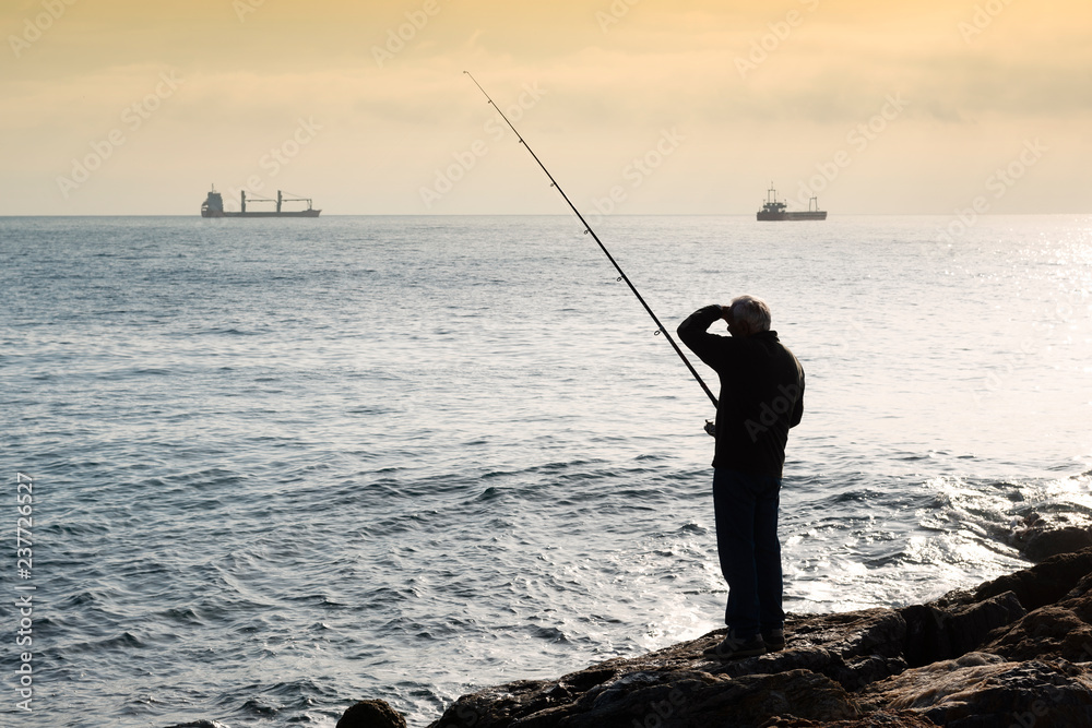 A fisherman standing with the fishing rod in his hand, he is on the rocks at sunset with his hand over his eyes to observe the sea, on the bottom an oil tanker and a barge sail on the horizon, the sky