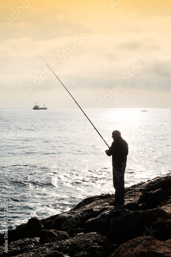 A fisherman standing with the fishing rod in his hand, he is on the rocks at sunset, on the bottom an oil tanker sails on the horizon, the sky is cloudy and golden.