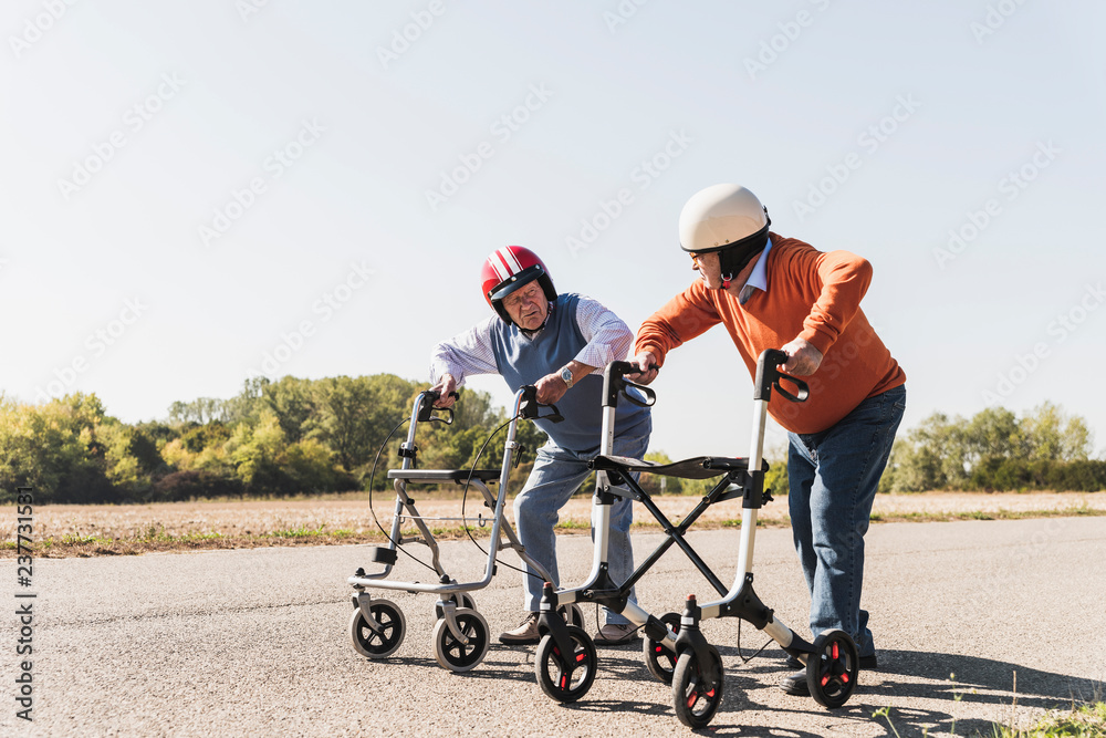Two old friends wearing safety helmets, competing in a wheeled walker race Two old friends wearing safety helmets, competing in a wheeled walker race