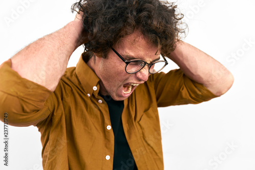 Angry handsome man with curly hair opens mouth widely, screams loud with hands on head, wears round glasses posing on againsr white wall. Copy space for your advertisement. People and negative emotion photo