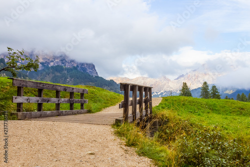 Small wooden bridge on a tourist pathway on a green hilly meadow with high mountains and cloudy sky in the backdround in Dolomites in Italy