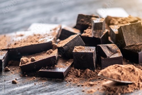 Pieces of dark bitter chocolate with cocoa powder on dark wooden background. Concept of confectionery ingredients