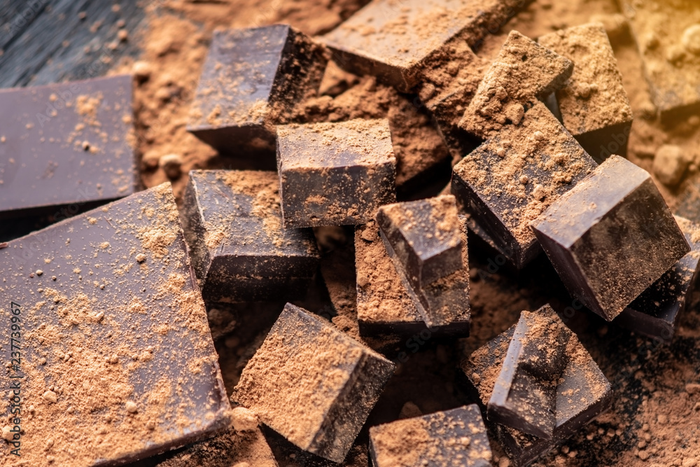 Pieces of dark bitter chocolate with cocoa powder on dark wooden background. Concept of confectionery ingredients