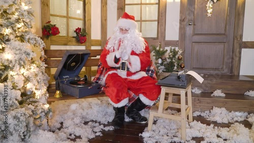 Santa Claus is printing the letter by typewriter, sits outside the house between christmas trees, drinks milk, listens music on vinyl by gramophone, preparing for Christmas