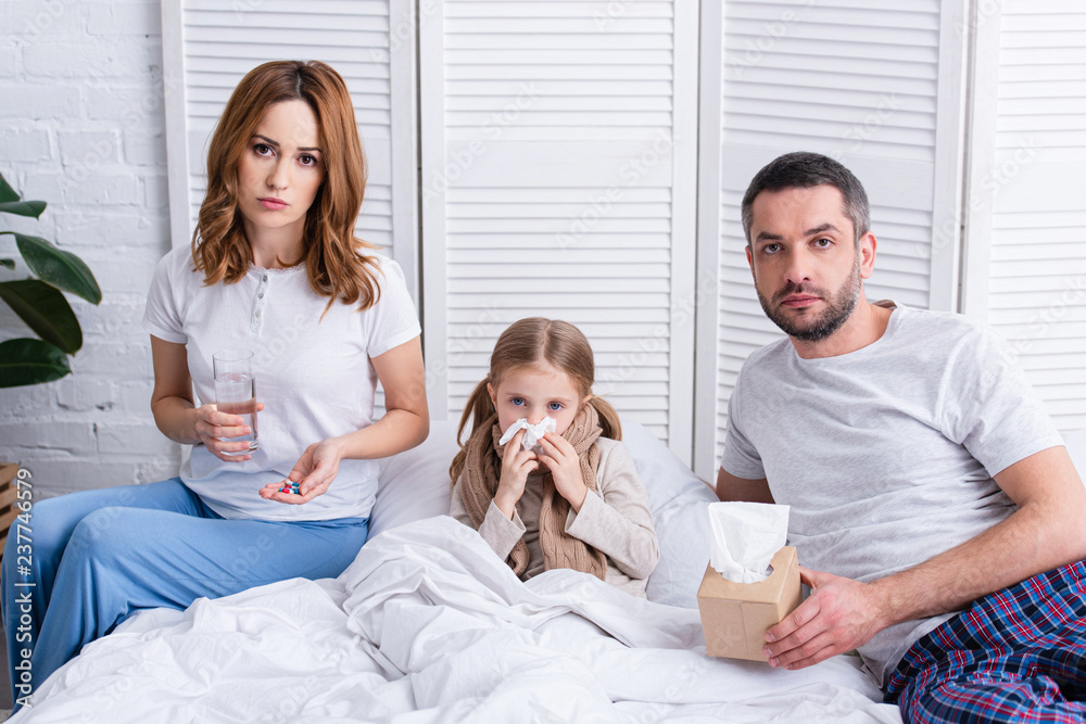 Obraz premium parents taking care of sick daughter in bedroom, giving her pills and glass of water, looking at camera