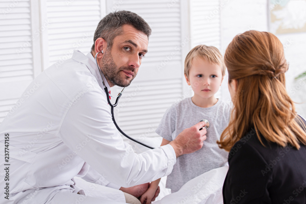 pediatrist in white coat examining sick boy with stethoscope and looking at camera at home pediatrist in white coat examining sick boy with stethoscope and looking at camera at home