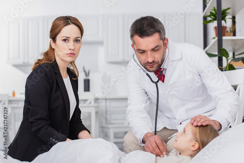 pediatrist in white coat examining sick kid with stethoscope at home, mother looking at camera