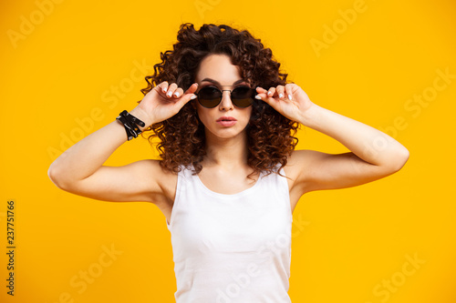 Young model expressing emotions while posing on indoor photoshoot. Stylish curly woman having fun in yellow studio