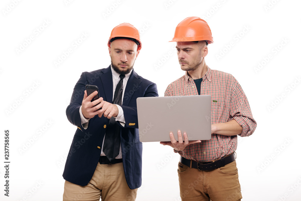 Portrait of two builders in protective orange helmets standing on white isolated background and looking at laptop display. Discuss construction project Portrait of two builders in protective orange helmets standing on white isolated background and looking at laptop display. Discuss construction project