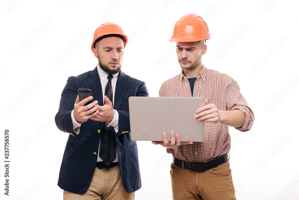 Portrait of two builders in protective orange helmets standing on white isolated background and looking at laptop display. Discuss construction project Portrait of two builders in protective orange helmets standing on white isolated background and looking at laptop display. Discuss construction project