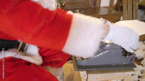 Santa Claus is printing the letter by typewriter, sits outside the house between christmas trees, drinks milk, listens music on vinyl by gramophone, preparing for Christmas