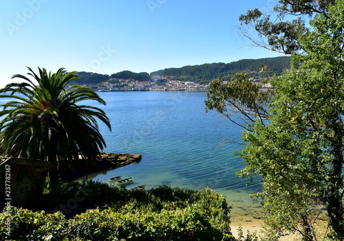 Small beach in a bay with clear water, trees, vegetation and boats. Sea with green and turquoise colours, sunny day. Coastal village, Galicia, Spain. 