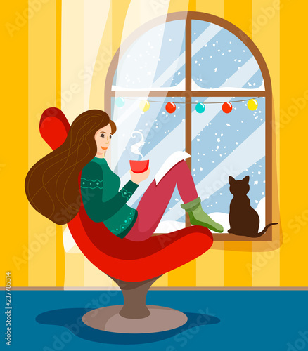 A girl sits by a winter garland-decorated window in an armchair - a chair with a hot mug of drink and reads a book, and a cat sits on a window sill with a snow over a snowball