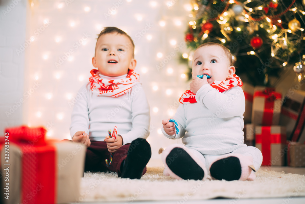 small children near the Christmas tree and fireplace with colored candies in their hands small children near the Christmas tree and fireplace with colored candies in their hands