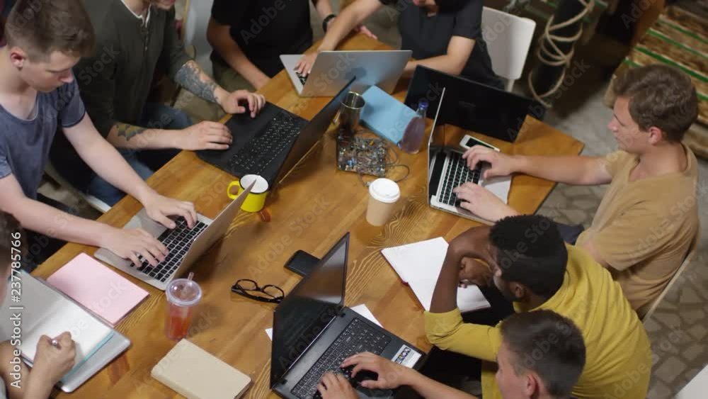 High angle view of multiethnic group of young people sitting at desk and using laptop computer and notebooks, camera rotating around axis