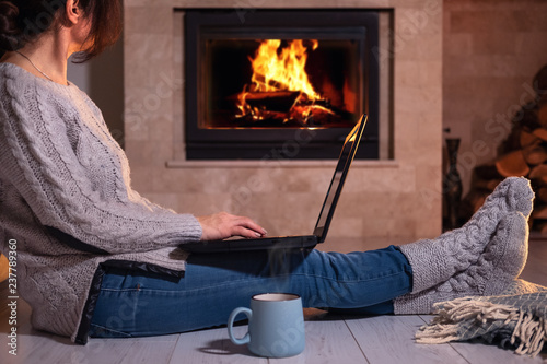 Young freelancer woman sits at the floor with a laptop on the fireplace background. 