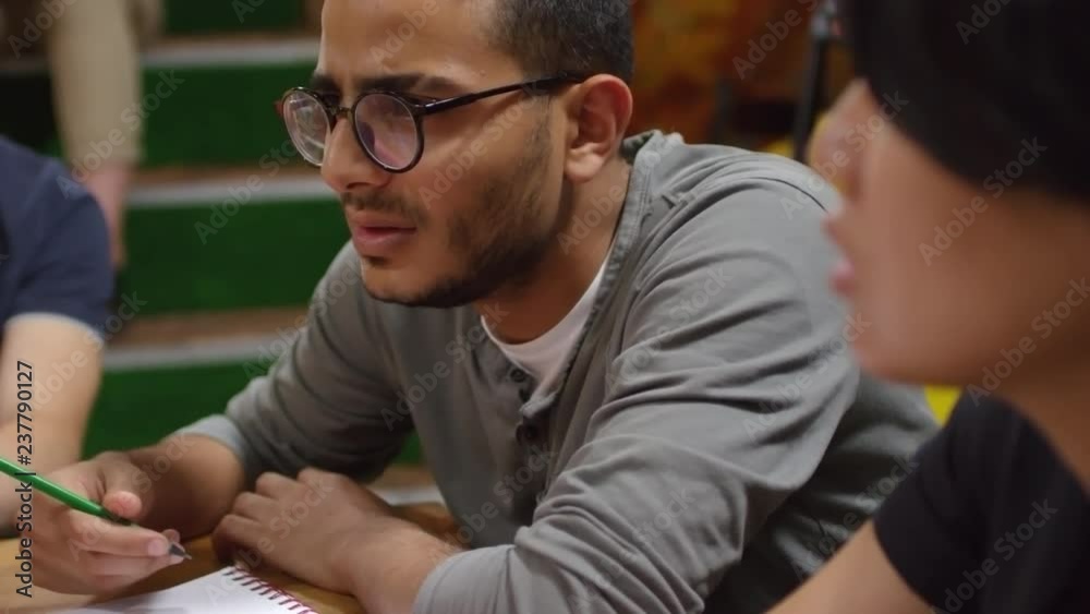 Medium shot of concentrated young Asian man in glasses sitting at desk and discussing task with groupmates during class or workshop Medium shot of concentrated young Asian man in glasses sitting at desk and discussing task with groupmates during class or workshop