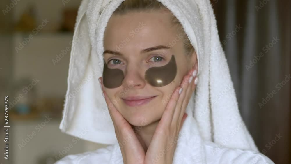 Young adorable woman in white towel turban doing her daily beauty care cosmetic routine, applies moisturizing patches in the bathroom and smiling at the camera. Young adorable woman in white towel turban doing her daily beauty care cosmetic routine, applies moisturizing patches in the bathroom and smiling at the camera.