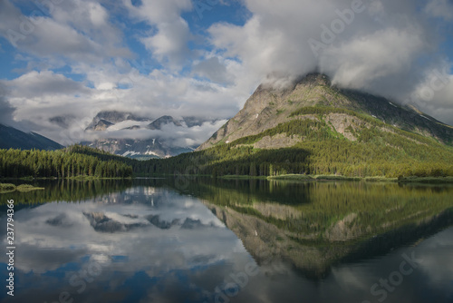 Billowing white clouds obscure the rugged peaks of the mountains surrounding Swiftcurrent Lake, which are perfectly reflected in the still water. photo