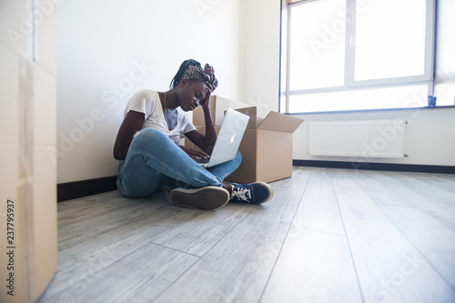 Sad young african woman sitting on floor near moving boxes and looking on screen of laptop. Young woman moving to new home. © F8  \ Suport Ukraine