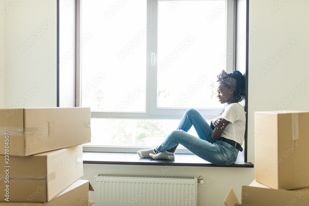 Portrait of young afro american woman moving in new home sitting on windowsill sourendet with cartoon boxes. Relocation concept, Portrait of young afro american woman moving in new home sitting on windowsill sourendet with cartoon boxes. Relocation concept,
