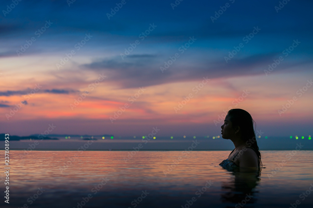 Black silhouette of asian woman on summer vacation holiday relaxing in infinity swimming pool with blue sea sunset view. Healthy happiness lifestyle