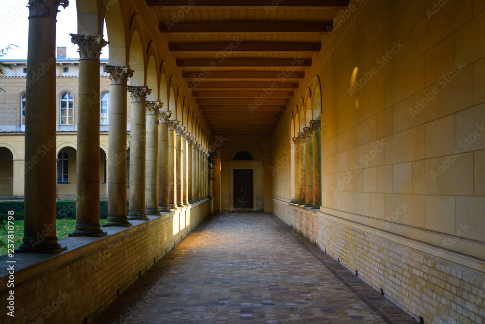 Diminish perspective view of inner corridor with yellow stone wall and arch columns frame windows around courtyard.