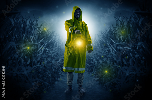 Man in raincoat at night coming from thicket and looking something with glowing lantern 