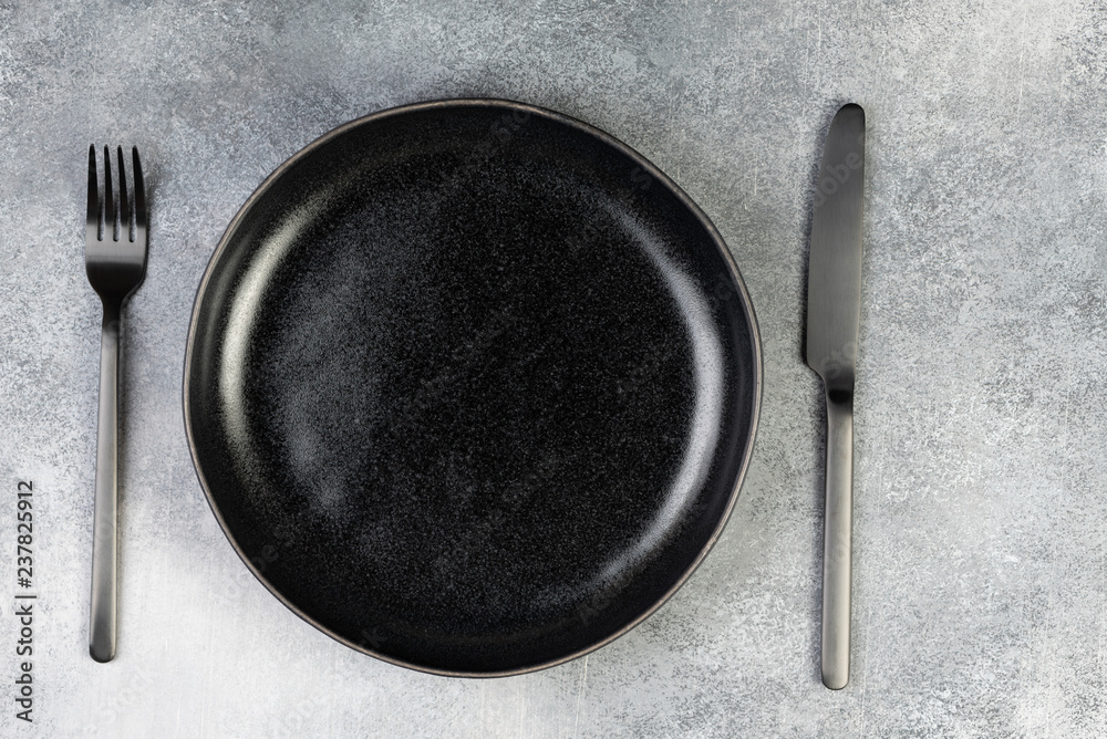 Empty black plate, fork, knife, spoon on blue background, top view