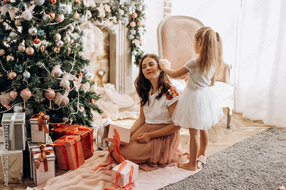 Happy young mother and her little daughter in nice dress sit near the New Year's tree and open New Year's gifts in the light cozy room Happy young mother and her little daughter in nice dress sit near the New Year's tree and open New Year's gifts in the light cozy room