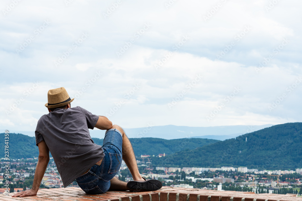 A young guy is sitting on the edge of a tall building and enjoying the view of the mountains and the city of Graz.