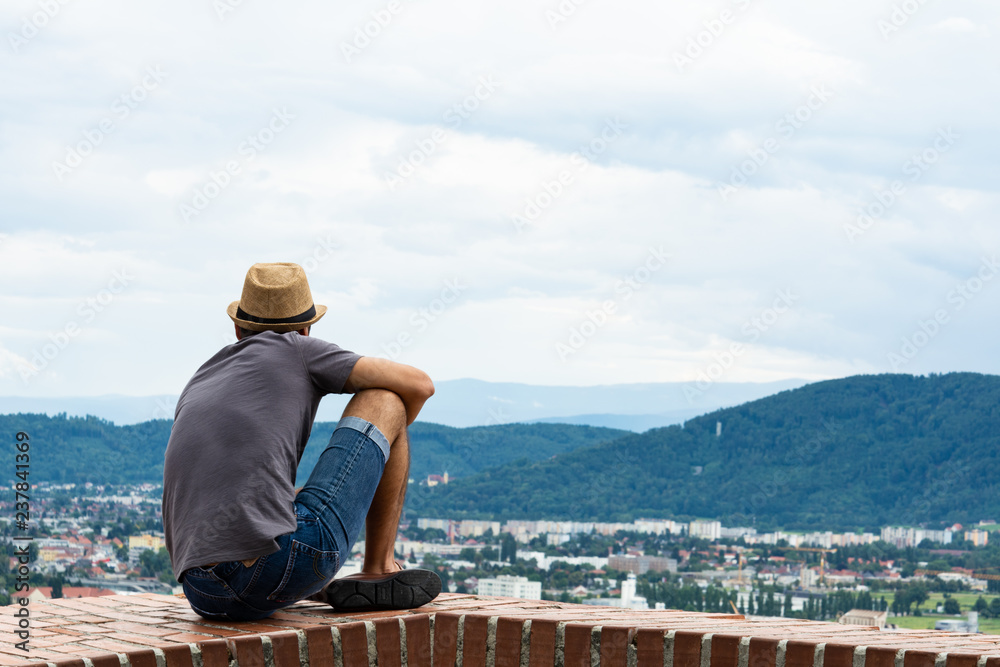 A young guy sit on the edge of a tall building and looks into the distance towards the mountains and the city of Graz.