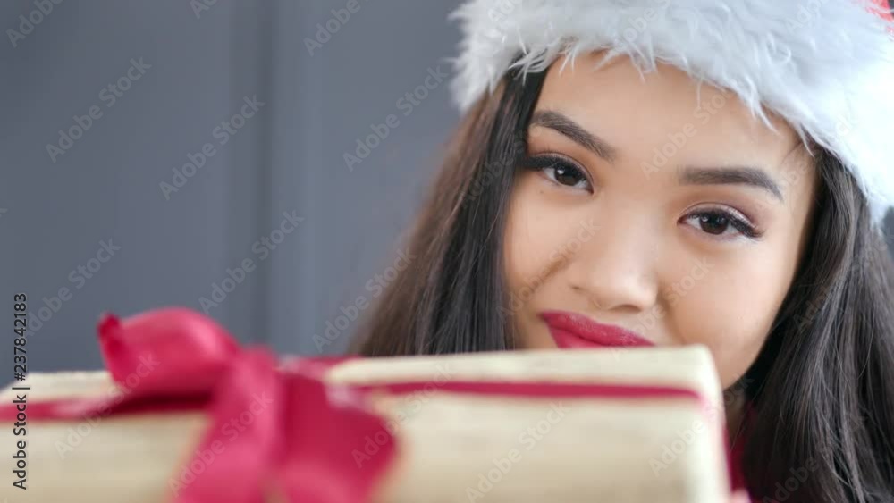 Close-up attractive young Asian Christmas girl wearing Santa Claus hat holding gift box with red bow Close-up attractive young Asian Christmas girl wearing Santa Claus hat holding gift box with red bow