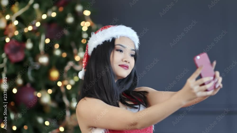 Smiling Asian young girl wearing Santa Claus suit taking selfie near Christmas tree using smartphone Smiling Asian young girl wearing Santa Claus suit taking selfie near Christmas tree using smartphone