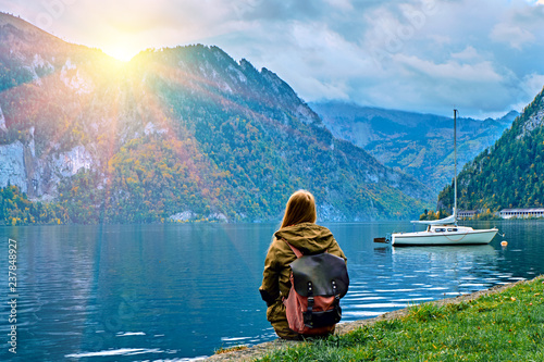 Young tourist girl with backpack looking at the beautiful scenic sunrise at Austrian alps lake. Hipster travel vacation in alps mountains concept. Nice yacht boats on calm peaceful alps lake