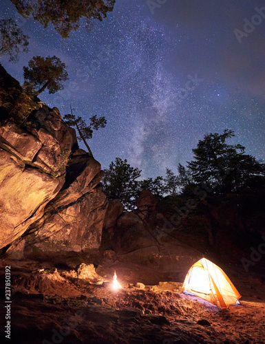 Mountain campsite at summer evening amid huge steep rock formation. Small tourist tent brightly lit by burning campfire under clear dark starry sky. Tourism  climbing  hiking and traveling concept.