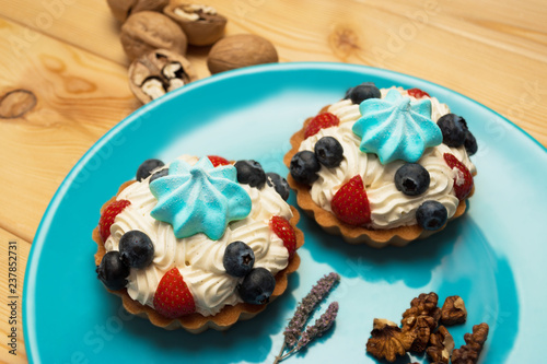 Two tartlets with white butter cream, blue marshmallow on top and strawberries and blueberries on blue plate and with walnuts and mint flower. The plate is on a wooden surface