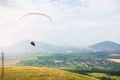 Professional paraglider in a cocoon suit flies high above the ground against the sky and fields with mountains