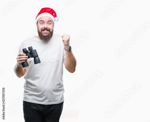 Young caucasian hipster man wearing christmas hat looking though binoculars over isolated background screaming proud and celebrating victory and success very excited, cheering emotion