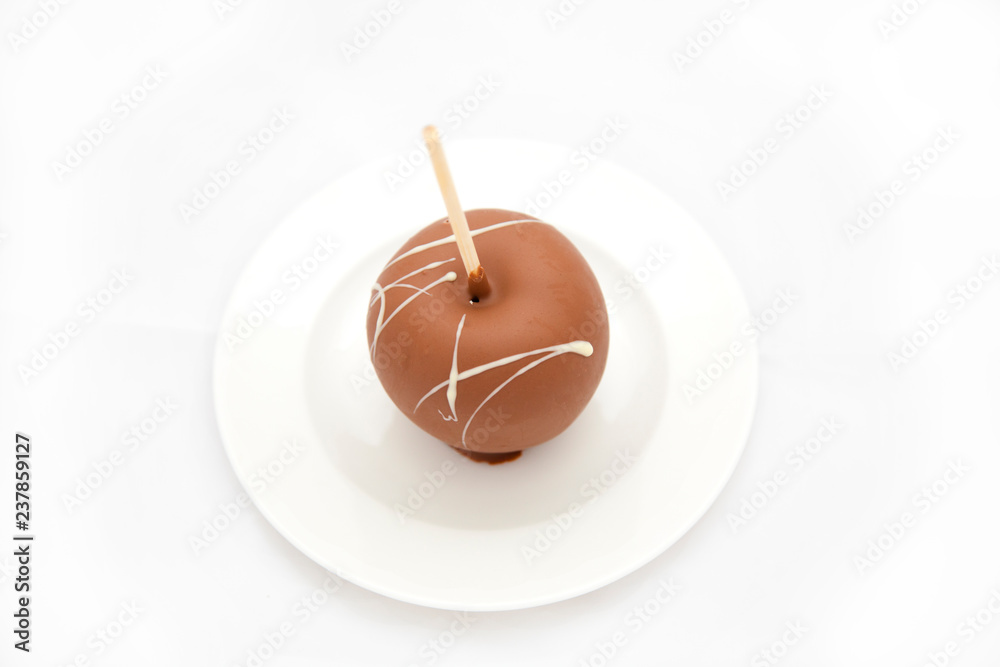 An apple covered with milk chocolate on a stick lies on a white plate. Christmas dessert on a white background. Isolated An apple covered with milk chocolate on a stick lies on a white plate. Christmas dessert on a white background. Isolated