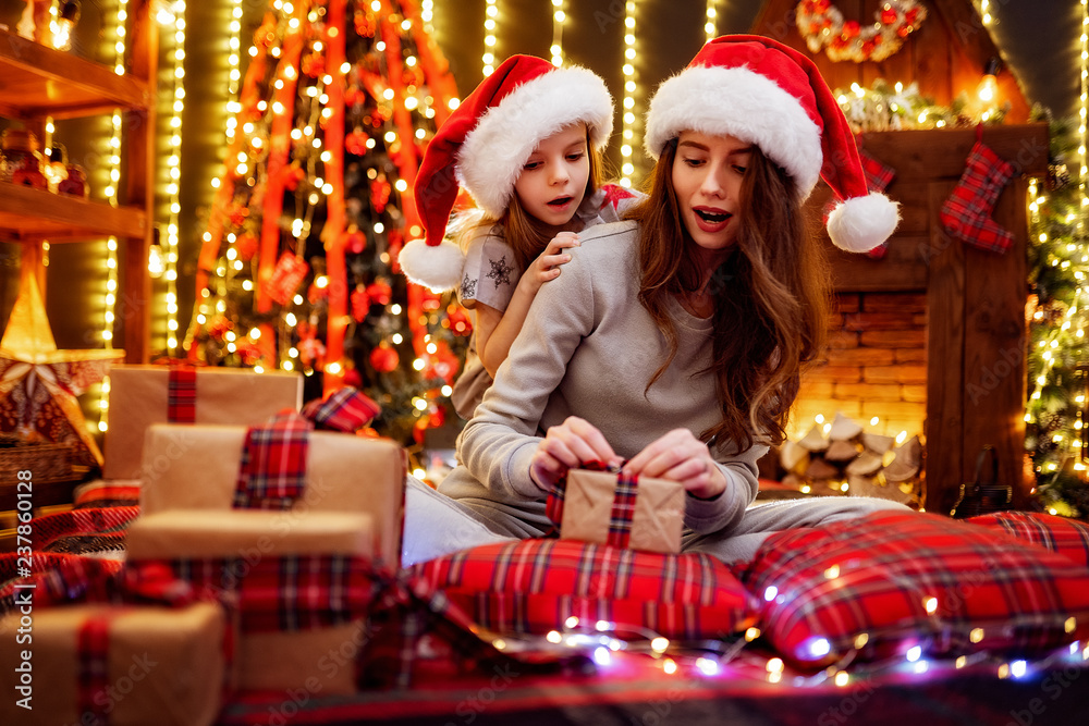 Cheerful mom and her cute daughter girl in santas hats and pajamas exchanging gifts. Parent and little children having fun near tree indoors. Loving family with presents in christmas room.