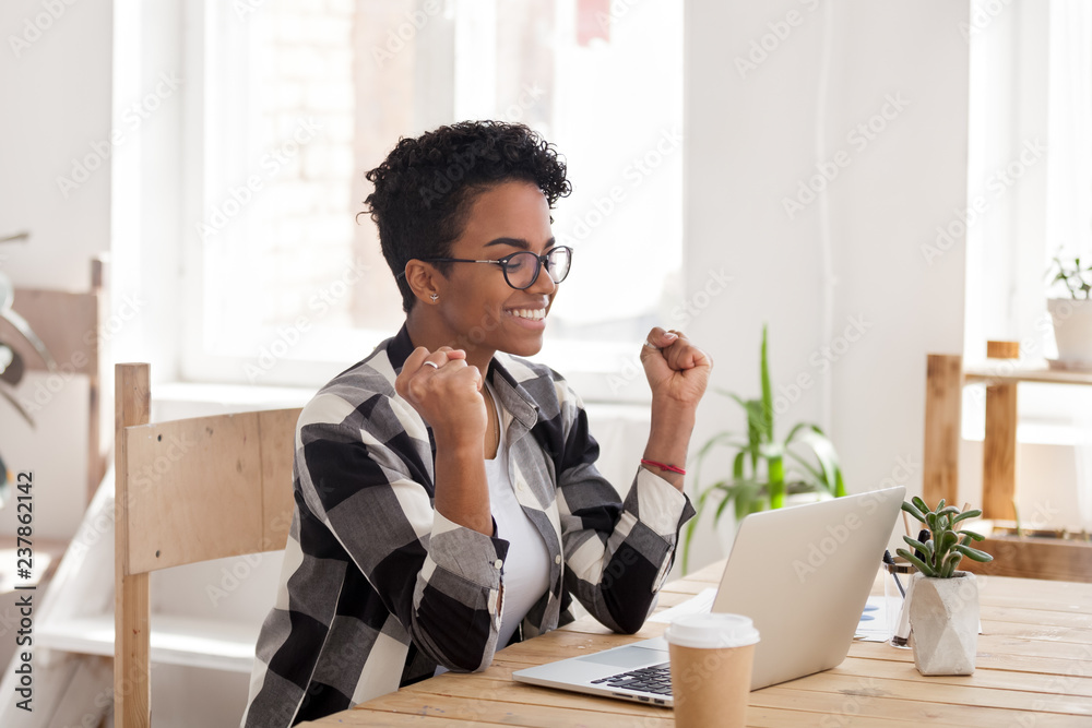 Excited African American woman in glasses read good news online at laptop, happy black female get promotional letter or email celebrating goal achievement, smiling girl look at computer lucky with win - obrazy, fototapety, plakaty 