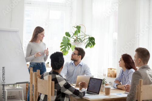 Millennial female employee make flipchart presentation for diverse employees during meeting, young mentor or coach talk giving educational business training to multiethnic workers in office